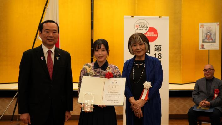 漫畫家左萱（中）手持獎狀及獎座，與外務省副大臣藤井比早之（左）、審查委員長里中滿智子（右）合影（台灣文化中心提供）