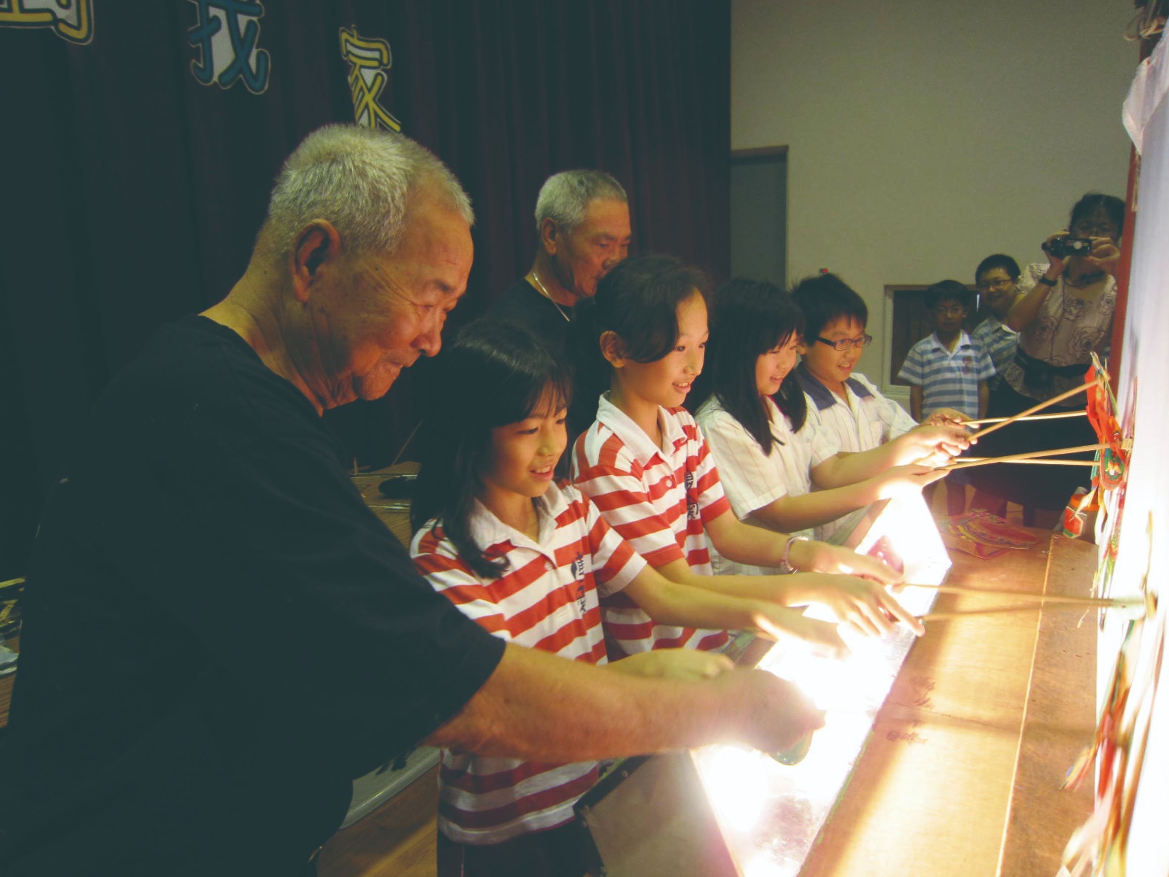 永興樂第三代張歲（左後一）昔日在自家影窗教導晚輩，如今與第四代張新國（左後二）在校園牽起無數孩子的手；對傳承百年的永興樂而言，那陣陣純粹的笑聲，就是老劇團最想留給未來的風景。（永興樂皮影劇團提供）