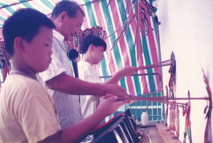 現任團長張信鴻（左一）幼年時，跟著阿公（張歲／左二）習藝；如今長大成為劇團接班人，帶著阿公進入校園傳遞家族五代志業。（永興樂皮影劇團提供）