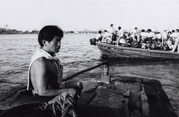 《走拍臺灣》04︰〈高雄旗津〉 1987年，旗津舢舨船上。 旗津原為高雄市西南部的沙洲半島，與鼓山、前鎮、小港等地隔高雄港相望。1967年為興建高雄港第二港口，截斷與臺灣本島相連處而形成島。1981年始興建過港隧道，1984年完工。由於過港隧道兩端均不在市區，居民長年以來，均以渡輪或舢舨船通勤往返旗津與鼓山之間。