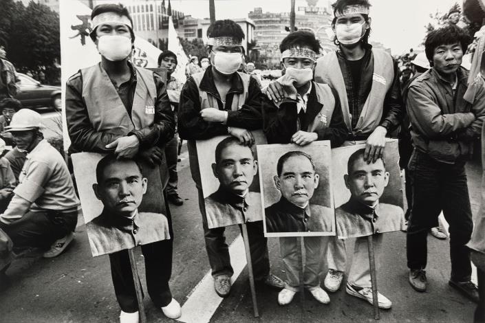 〈還我客家語遊行〉 1988年，立法院前。 本作品為1988年客家族群發起還我客語運動遊行，遊行隊伍行至立法院前，要求立法還予客家族群權益。畫面中客家族群帶著孫文肖像，強調國父亦為客家人，應尊重國父的母語文化。