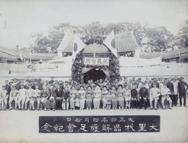 臺中州廳長官私人寫真帖（二）55 大里杙街在今臺中市東南部，1915年4月該街放足者已達1,200人，未放足者僅餘152人。大里杙解纏足會乃於4月10日上午11時集合已放足會員300人，在大里杙區役場前舉行解纏足祝賀會，會後舉行餐會，並贈送出席會員木屐一雙。本照片係祝賀會結束後所拍攝紀念照，照片中臨時搭建牌坊上方寫著「祝我自然」四字。