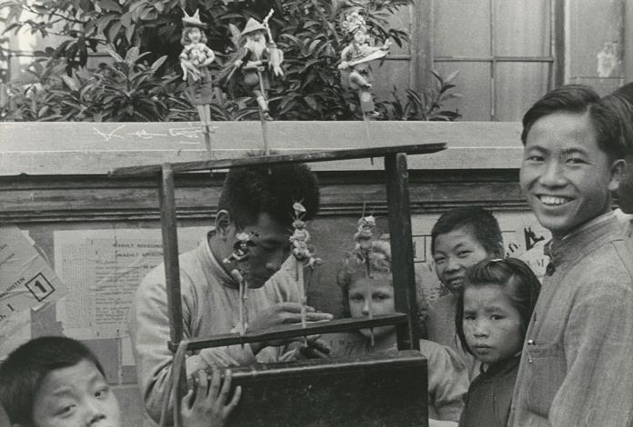 〈捏麵人〉 位在照片中央的是捏麵人展示的木架子，架上的捏麵人偶有手持大刀、頭戴盔帽的豫劇武生，還有身披簑衣、頭戴笠帽和腰間繫只葫蘆的老叟，最特別的是一尊穿著及膝裙套裝，頭戴女士帽及抱著寵物的西方女性。一群孩童圍繞在師傅四周，年齡大小不一，除了華人面孔，還有一位外國女孩。