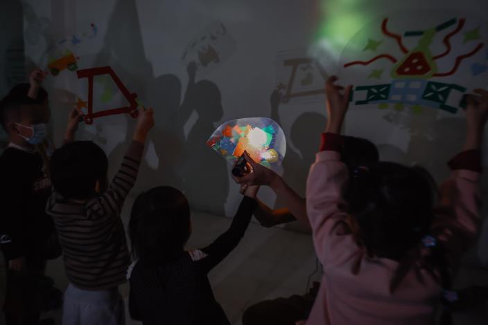 10光與影的紅綠燈派對：孩子拿著完成的車子戲偶，在光影情境中模擬交通情境。透過「停、看、聽」等動作，讓孩子在遊戲中理解不同顏色燈號所代表的行動。（此圖為綠燈）